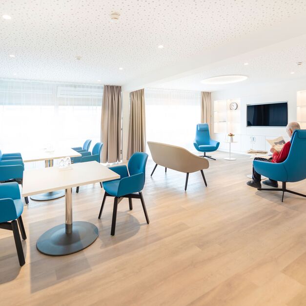 Heller Aufenthaltsbereich mit Tischen, Stühlen und Flat-TV. Ein Patient sitzt auf einem Sessel und liest Zeitung.
